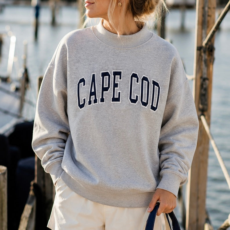 A woman wearing a light gray crewneck sweatshirt with 'CAPE COD' printed in navy blue varsity letters across the chest, standing outdoors near water and wooden dock structures.