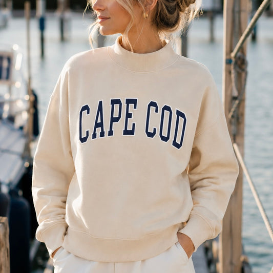 A woman wearing a cream-colored mock neck sweatshirt with 'CAPE COD' printed in navy blue varsity lettering across the chest, standing outdoors near water and boats.
