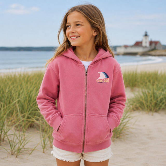 A young girl stands on a sandy beach wearing a pink zip-up hoodie with a small 'Cape Cod' logo on the chest, paired with white shorts. The background features beach grass, the ocean, and a lighthouse under a blue sky.