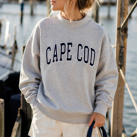 A woman wearing a light gray crewneck sweatshirt with 'CAPE COD' printed in navy blue varsity letters across the chest, standing outdoors near water and wooden dock structures.