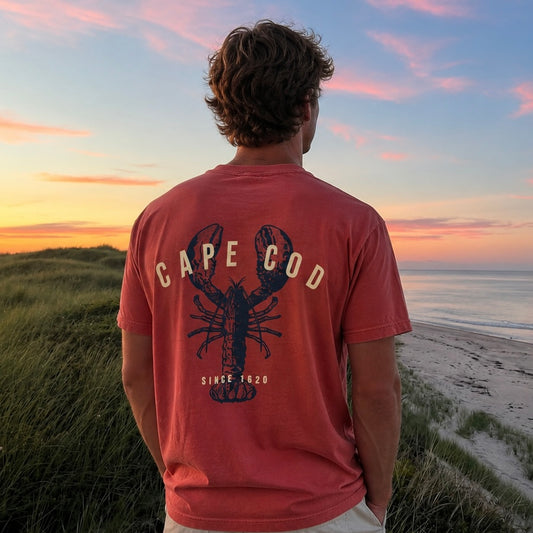 A man viewed from behind wearing a rust-orange t-shirt with a lobster graphic and 'CAPE COD' text, standing on grassy dunes overlooking a beach at sunset.