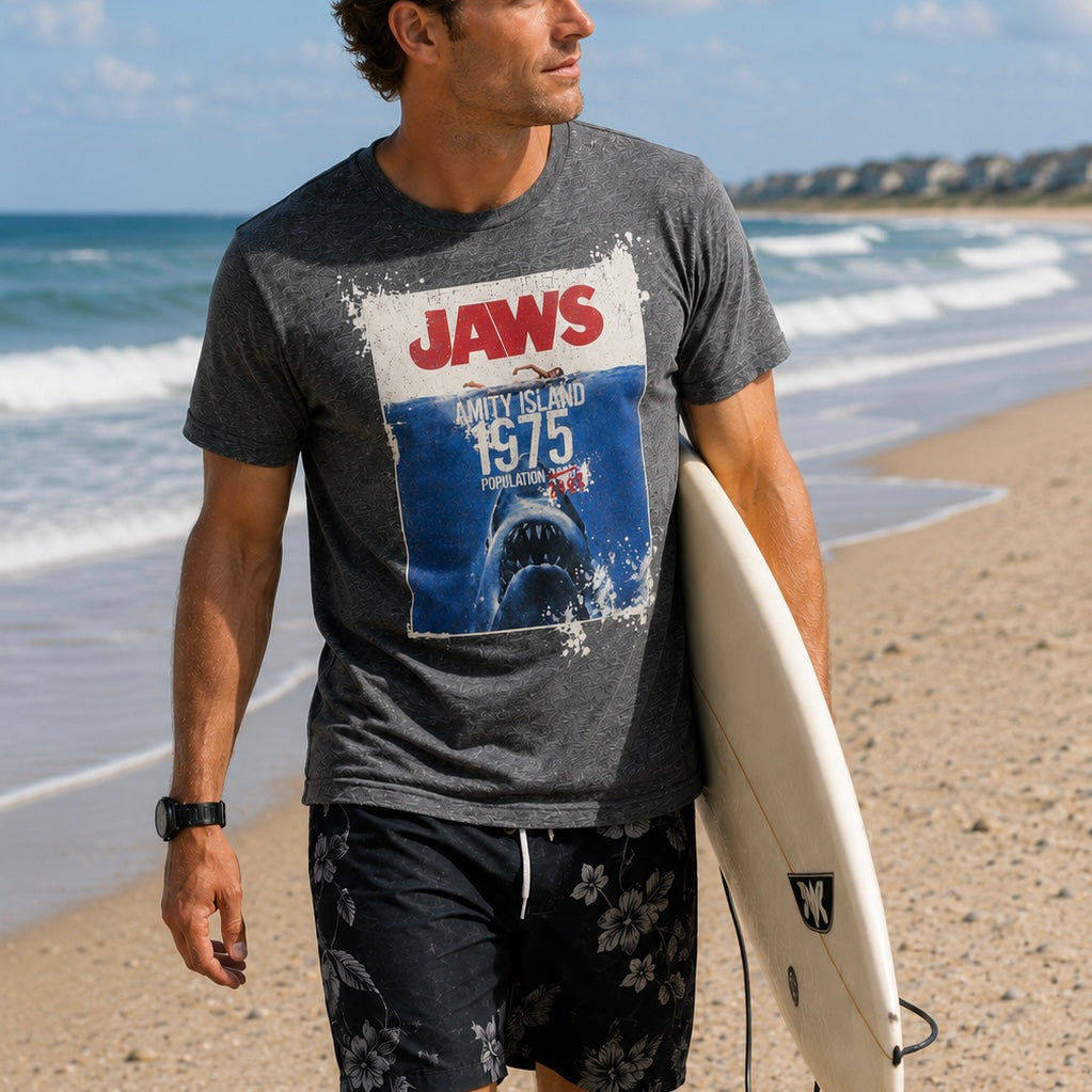 Man on a beach wearing a 'Jaws' t-shirt and holding a surfboard.