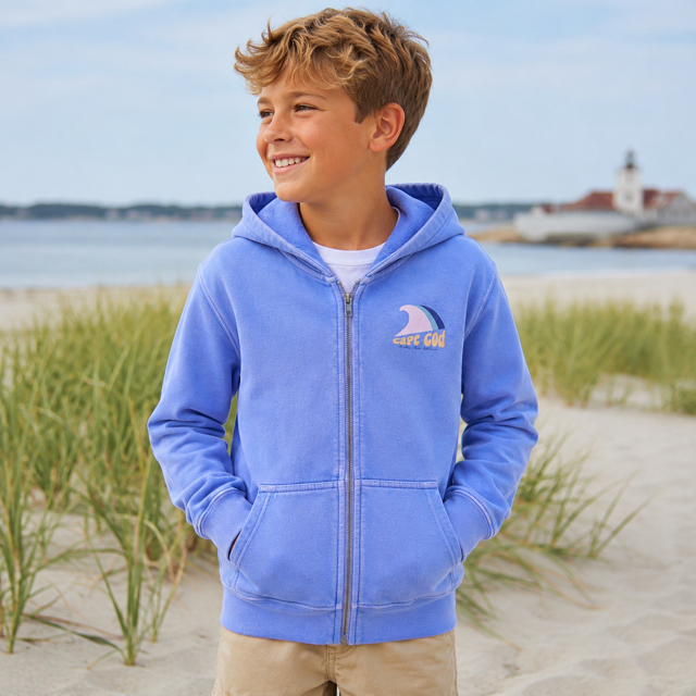 A young boy smiling on a beach dune wearing a light blue zip-up hoodie with a shark graphic and 'AFC Club' text on the chest, paired with khaki pants.